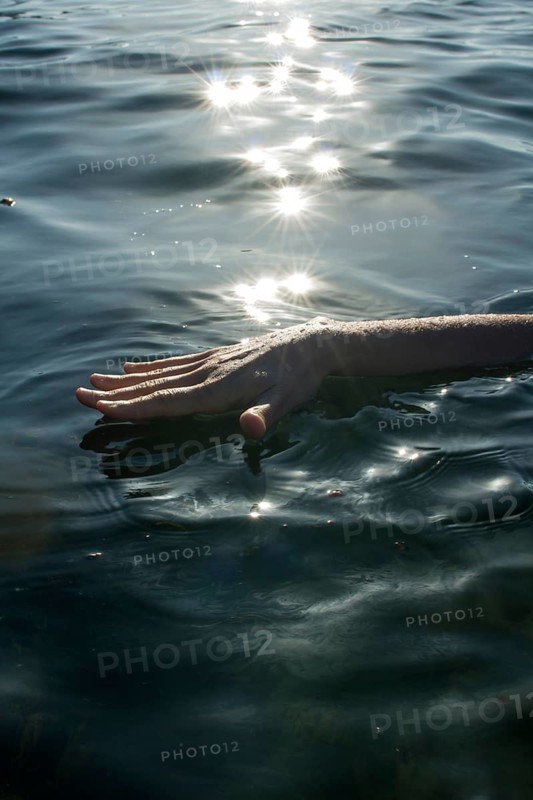A hand gently touches the surface of a tranquil water body, creating ripples. Sunlight sparkles on the water, enhancing the peaceful atmosphere of the scene