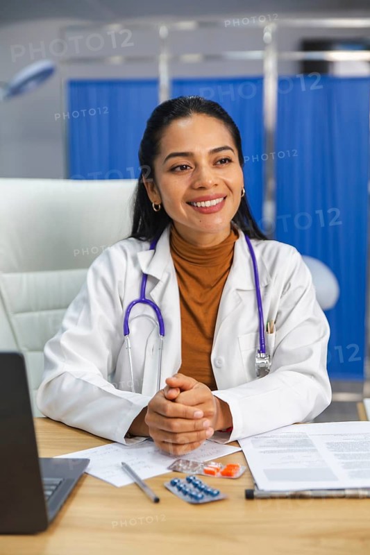 A smiling doctor with a stethoscope around her neck sits at a desk with paperwork and medication on display. The setting is a bright medical office environment