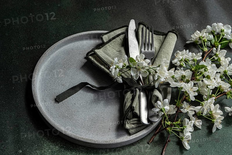 A sophisticated table setting features a gray plate with neatly arranged cutlery and a green napkin, adorned with delicate white blossoms for a fresh, springtime look
