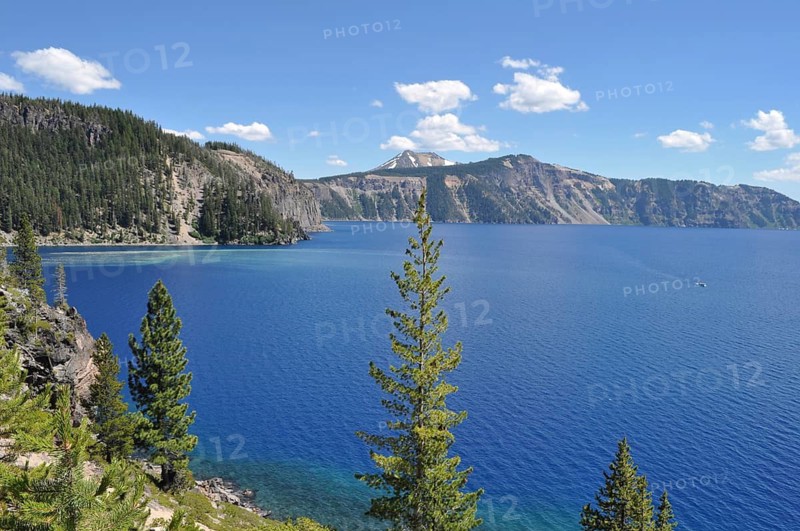 View of blue lake and green conifers against a mountainous backdrop under slightly cloudy sky, Crater Lake National Park, Oregon, USA