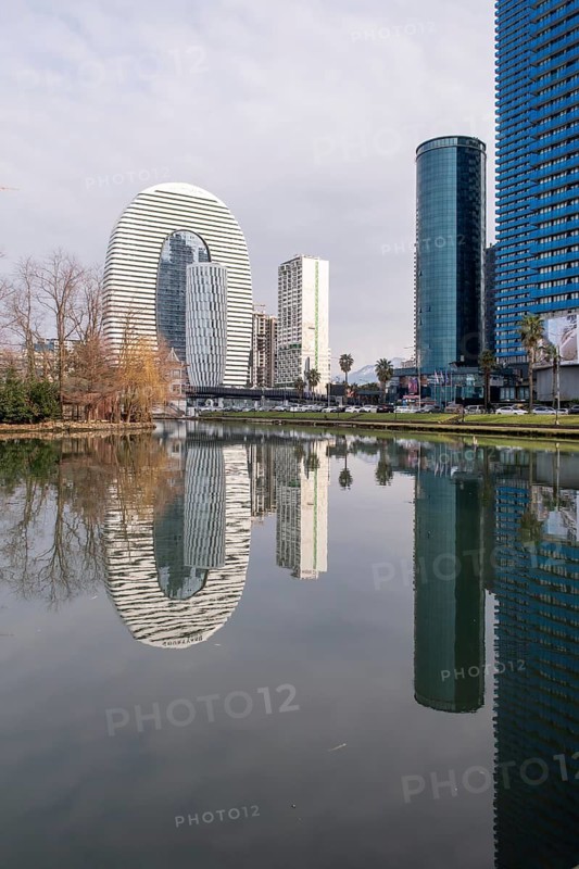 A stunning view of Batumi's modern skyline featuring distinctive architecture, mirrored in the tranquil waters of a nearby lake. Trees and clear skies enhance the scene's beauty