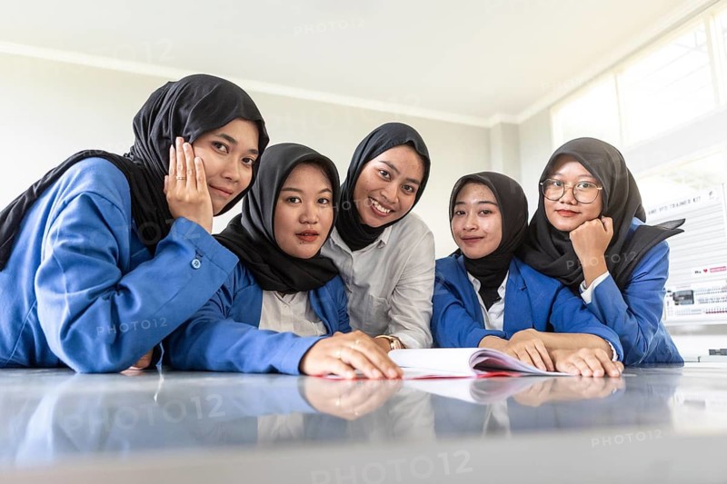 Group of Asian Muslim women sitting together in classroom, wearing hijabs and blue uniforms. They are smiling, displaying enthusiasm and camaraderie in their educational environment