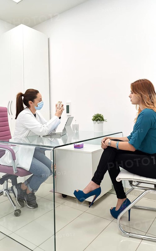 A dentist in a white coat and mask consults with a patient in a modern dental clinic. The office features sleek furniture and a calm, professional atmosphere