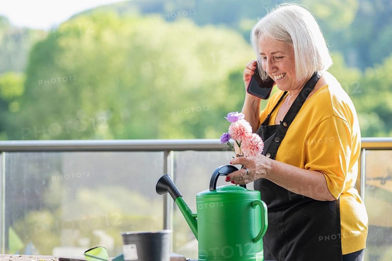 A cheerful elderly woman in a yellow shirt and apron holds colorful flowers while speaking on her phone. She stands by a green watering can, surrounded by nature