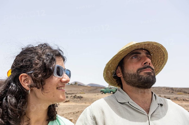 A blind man and woman stand together in a desert terrain. The man is wearing a straw hat and an old vehicle is seen in the background, symbolizing adventure and connection