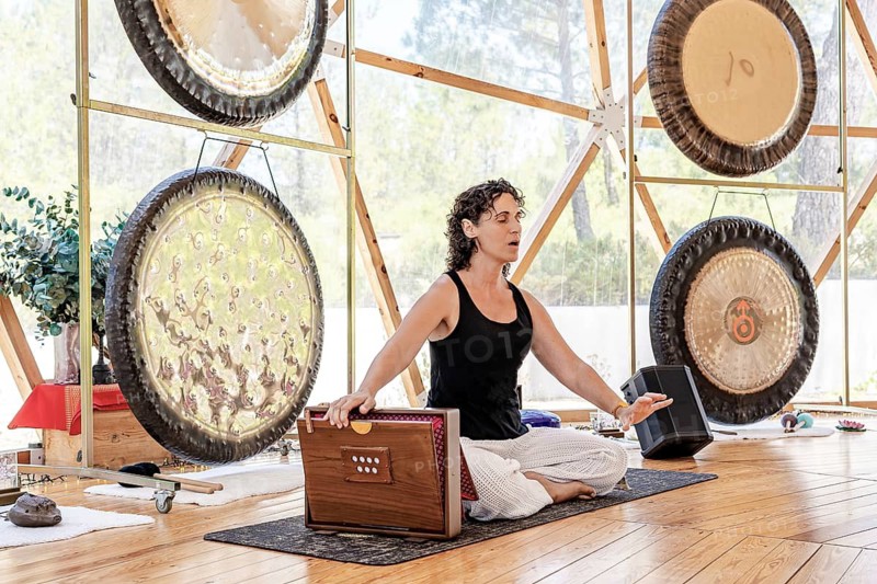 A female sound therapist plays a shruti box with her eyes closed, emphasizing tranquility. Gongs surround her, enhancing the holistic atmosphere in the wellness retreat