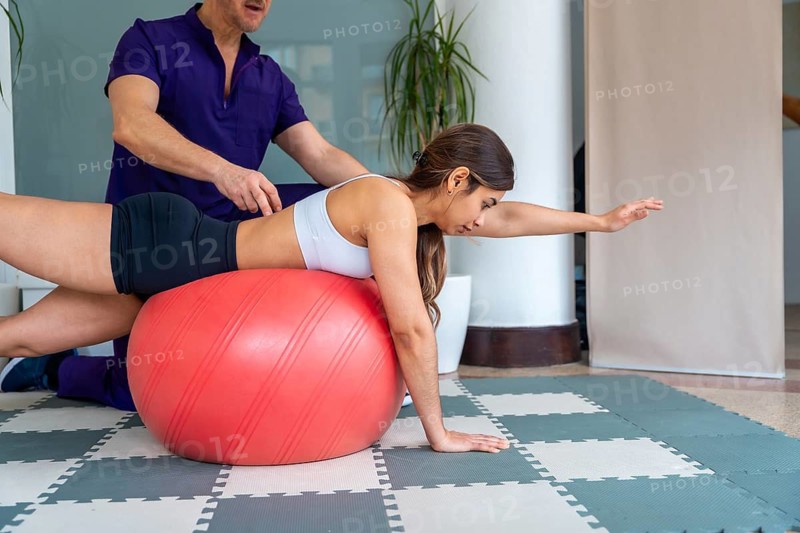 Physiotherapist assisting a young woman balancing on an exercise ball, performing core strength and stability exercises for medical recovery and physical rehabilitation therapy