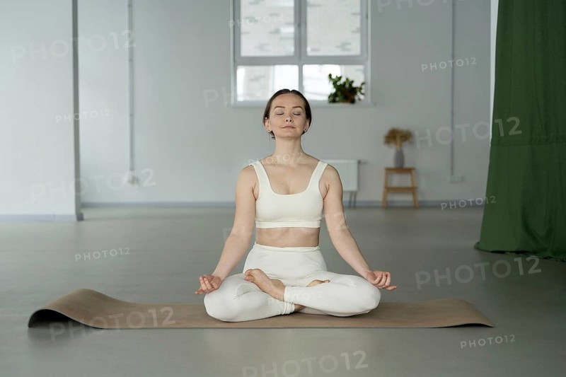 A woman practices meditation in a serene studio. She seated cross legged on a yoga mat, eyes closed, wearing a light outfit that matches the calm, minimalist decor