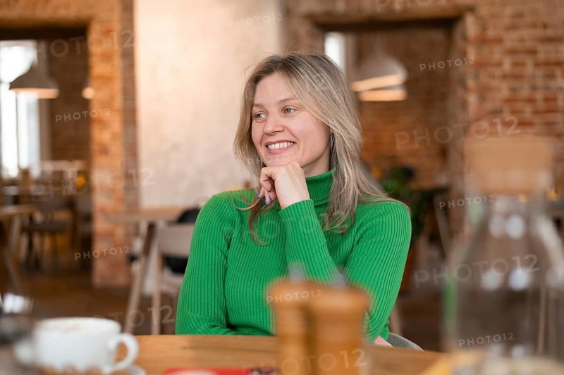 A woman in a green sweater smiles while sitting at a cafe table. The atmosphere is warm and inviting, with coffee and food items subtly visible, creating a cozy breakfast scene