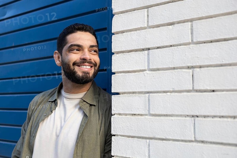 A man with a beard smiles while leaning against a white brick wall of the city, confidently standing next to a door blue. The lighting highlights his content expression