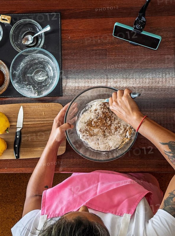 An overhead view of a woman mixing cookie dough in a glass bowl on a wooden countertop. Various kitchen utensils, ingredients, and a cutting board with lemons are visible