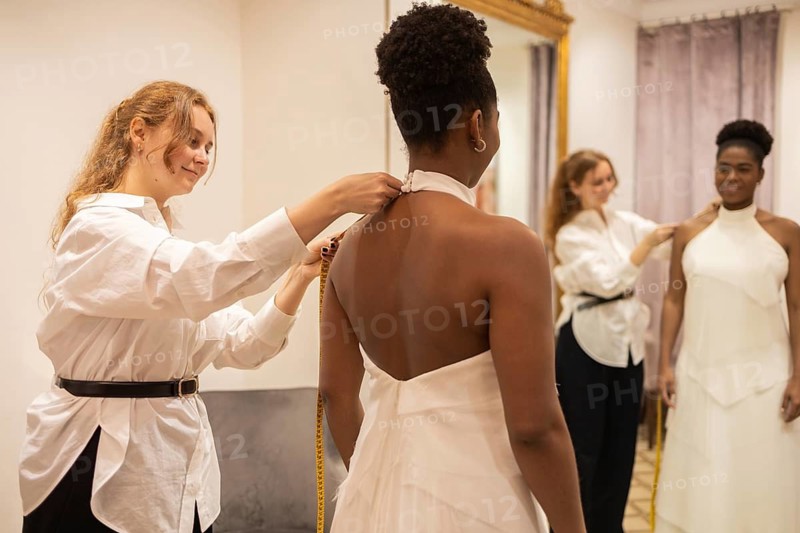 A seamstress adjusts a wedding dress for a bride in a fitting room. The bride stands in front of a mirror, admiring the elegant gown as final touches are made