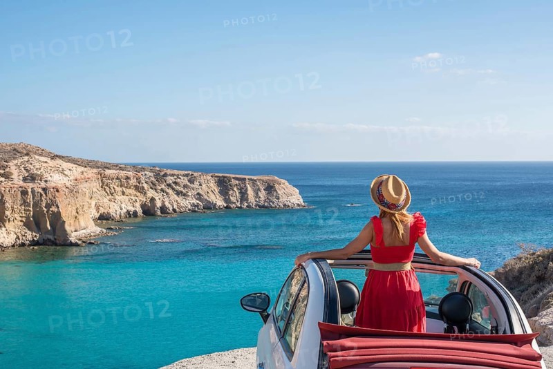 A woman in a red dress stands beside a convertible, overlooking the turquoise waters and rugged cliffs of Tsigrado Beach on Milos Island, Greece, under clear blue skies