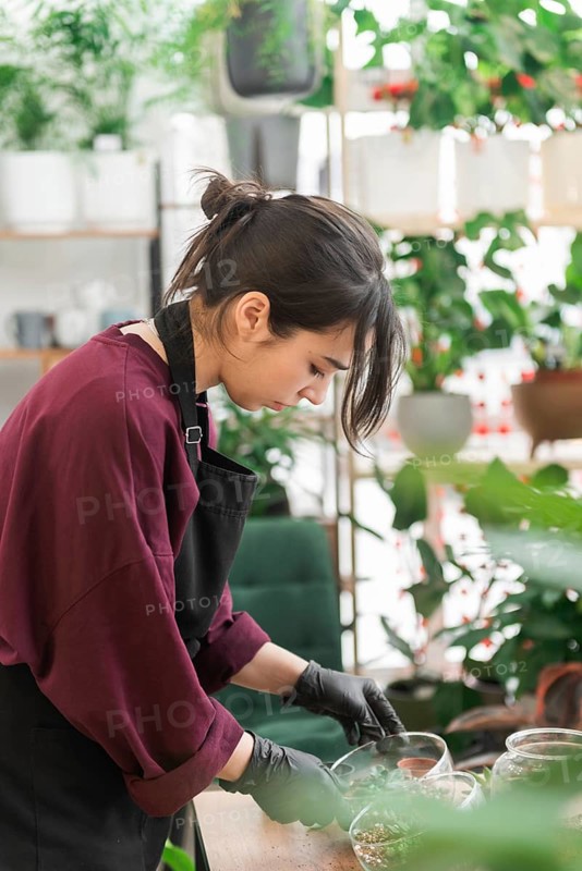 A woman wearing a black apron and gloves tends to plants in a nursery, surrounded by lush greenery. She appears focused and dedicated to her work