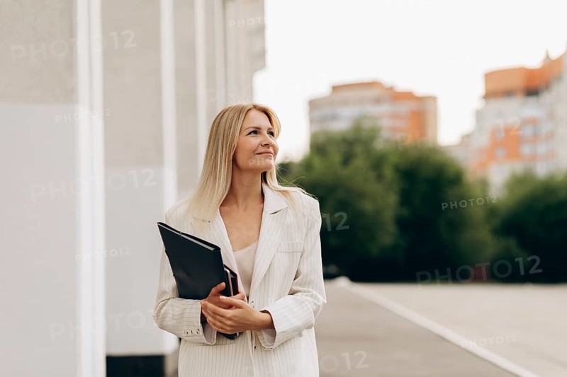 A professional woman in a light suit stands outside with confidence, holding a folder. Blurred cityscape and greenery in the background, conveying a sense of ambition