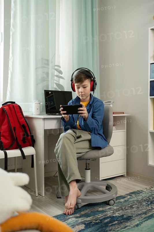 A young boy, wearing headphones, deeply focused on playing a game on his smartphone while sitting in a modern, bright room. A backpack and laptop are placed nearby
