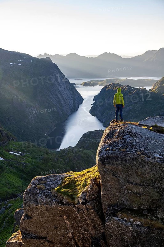 Mountaineer, young man standing on rocks and looking into the distance, at sunrise, fjord Trollfjord and mountains, hike to Trollfjord Hytta, at Trollfjord and Raftsund, Lofoten, Nordland, Norway
