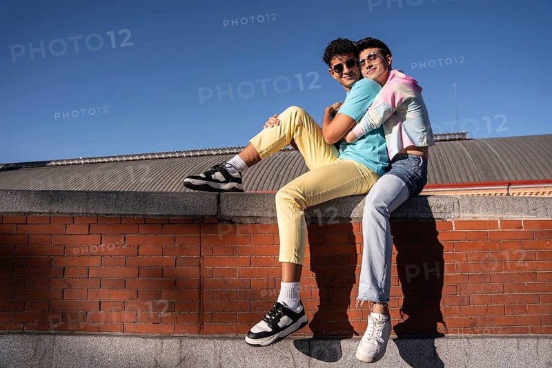 A joyful young gay couple sits on a brick wall, embracing and smiling under a clear blue sky. Their casual attire and sunglasses reflect a carefree, sunny day outdoors