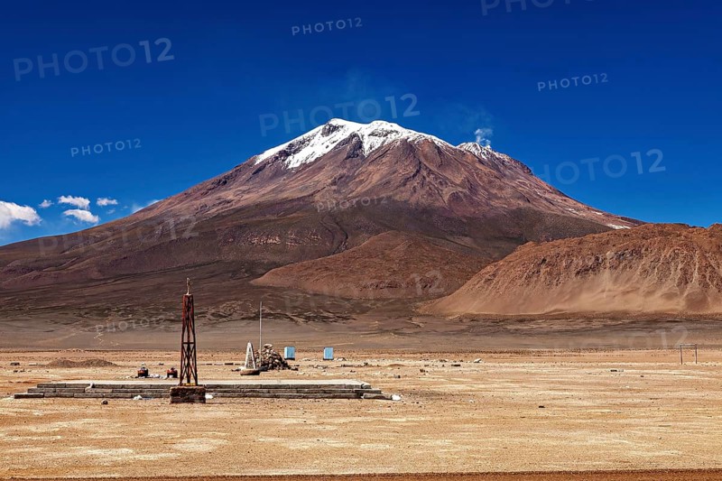 Isolated landscape with a large, snow-covered volcano and a small tower-like structure, the town of Ollagüe in the Chilean Atacama Desert with the Ollagüe volcano of the same name in the background