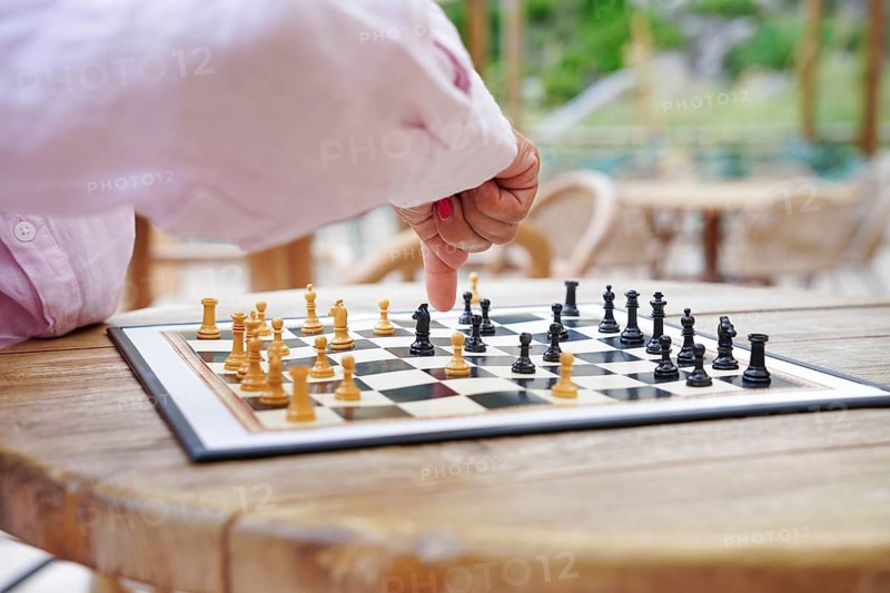 A person engaged in an outdoor chess game, showcasing strategic hand movement over a wooden table Natural light highlights the chess pieces, creating a tranquil scene