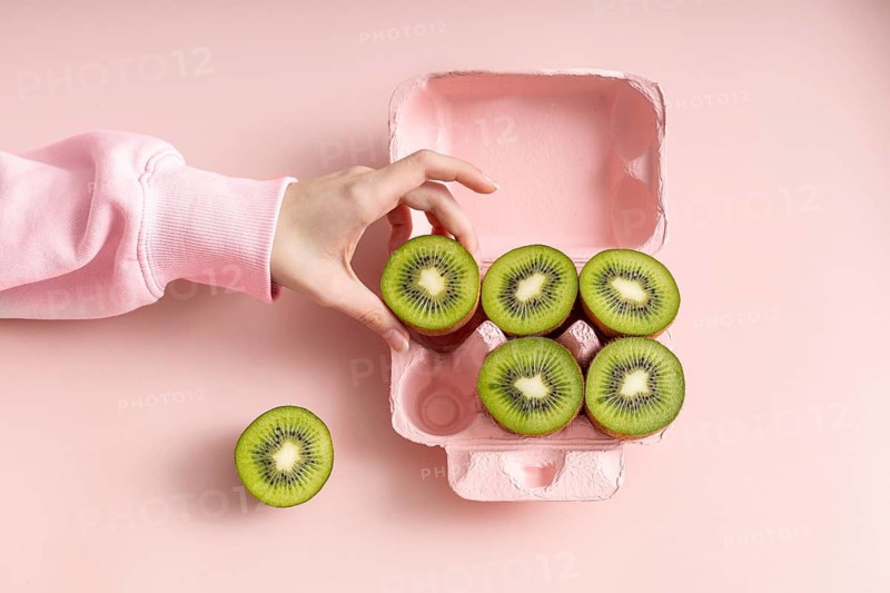 A cropped unrecognizable, hand adjusts a row of fresh kiwi slices neatly arranged in a pink egg carton, presenting an artistic and colorful healthy food concept
