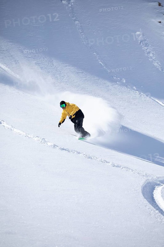 A snowboarder in a yellow jacket expertly navigates a snowy mountain slope, kicking up fresh powder as they descend. The bright snow contrasts with their colorful outfit