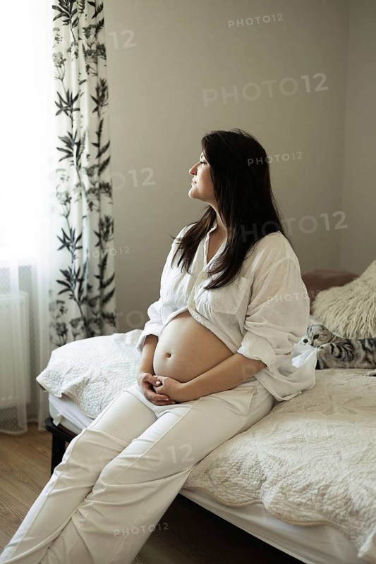 A serene pregnant woman sits on a bed, gazing thoughtfully, embodying the anticipation and preparation for motherhood. Her gentle posture reflects the beauty of expecting a baby