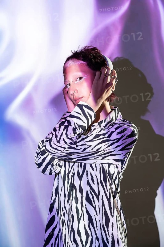 A young, trendy woman poses confidently in a zebra print shirt, illuminated by soft purple and blue lighting, exuding a modern, fashionable vibe with unique accessories