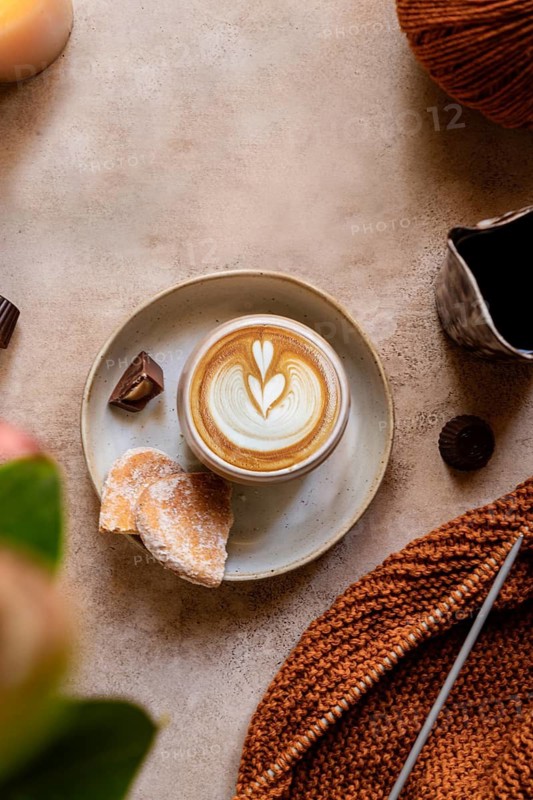 A warm scene featuring a knitted fabric beside a creamy latte art coffee, accompanied by cookies and chocolate, set on a rustic surface for a cozy autumn vibe