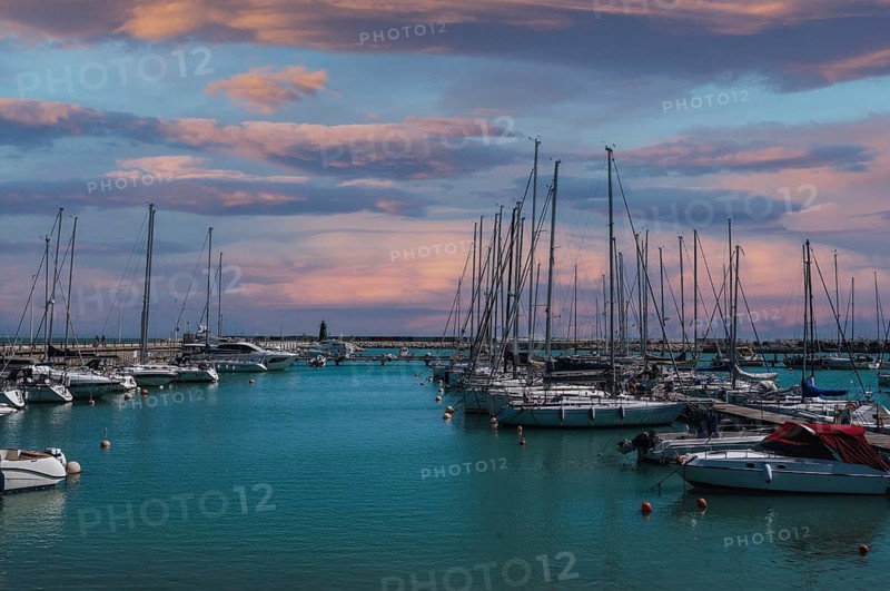 Sailboats gently bob in a serene harbor near Civitanova Marche, Italy. The sky is painted with warm sunset hues, casting a peaceful ambiance over the sea