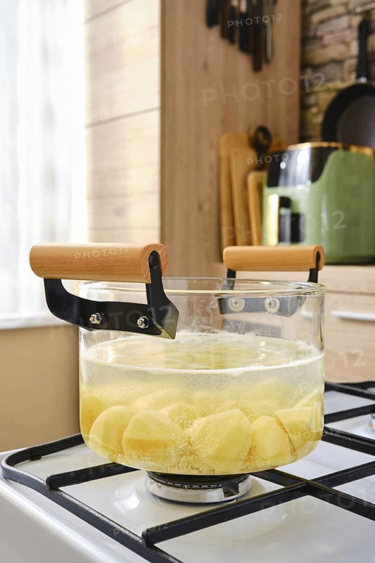 A glass pot filled with chopped potatoes simmers on a stove in a warm kitchen. The bright light illuminates the space, highlighting kitchen tools and utensils in the background