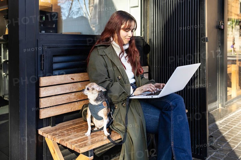 Woman seated on a wooden bench, typing on her laptop outside a cafe. Her small chihuahua sits beside her, creating a cozy, peaceful ambiance. Casual, urban scene captures leisure
