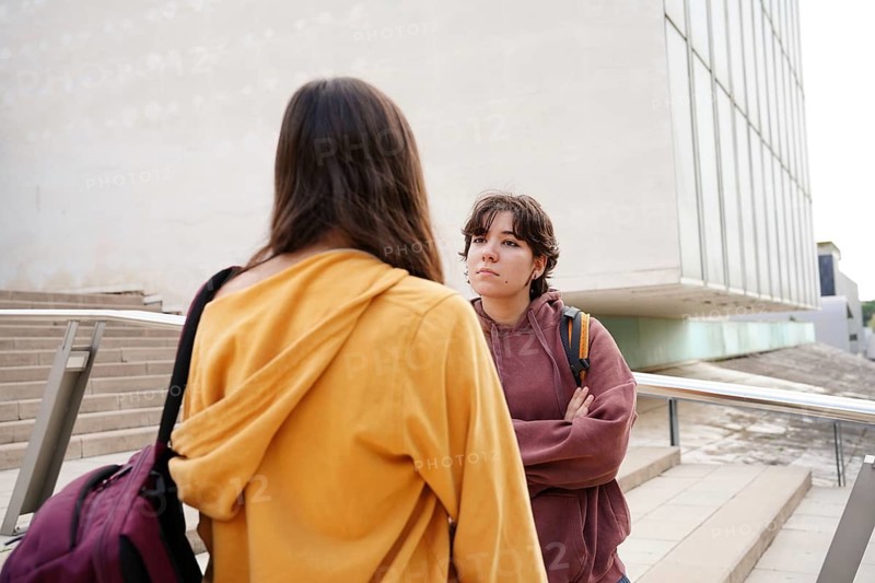 Two teenagers, a non-binary individual and a female, engage in conversation after returning to school, emphasizing the themes of friendship and education