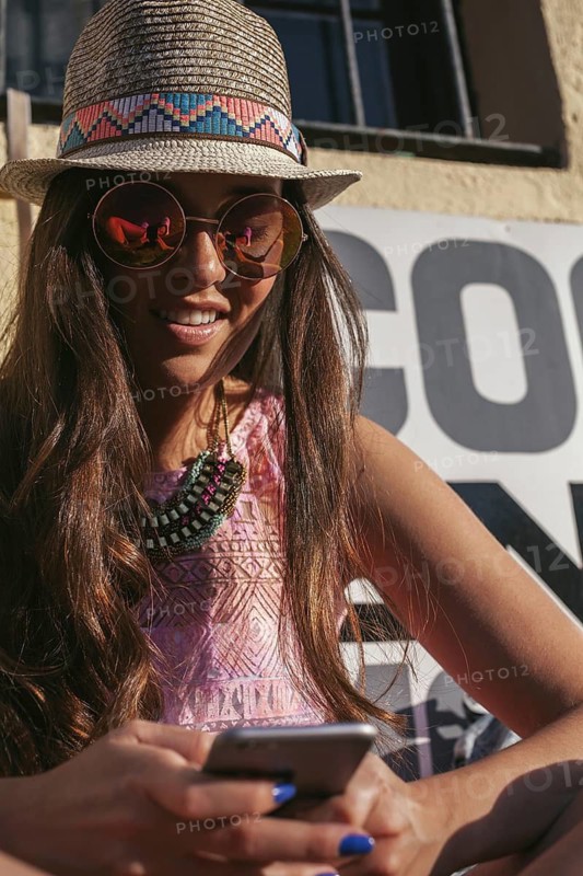 Stylish young woman in a summer hat and sunglasses, smiling while looking at her phone. She sports a colorful necklace and a vibrant outfit, embodying a trendy summer vibe
