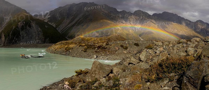 A vibrant rainbow arcs over Mount Cook in New Zealand during autumn. The scene captures the rugged mountain landscape, calm turquoise waters, and dynamic weather