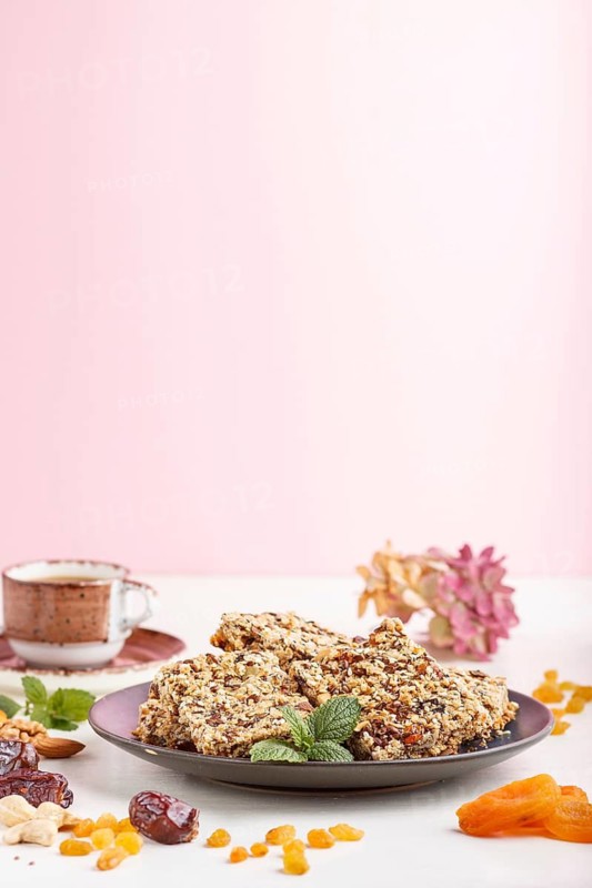 Homemade granola from oat flakes, dates, dried apricots, raisins, nuts in blue ceramic plate with a cup of coffee on a white and pink background. Side view, copy space