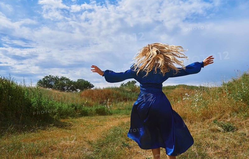 A joyful blond woman in a vibrant blue dress playfully twirls in a lush grassy field under a broad blue sky. Her carefree spirit and the natural setting evoke a sense of freedom