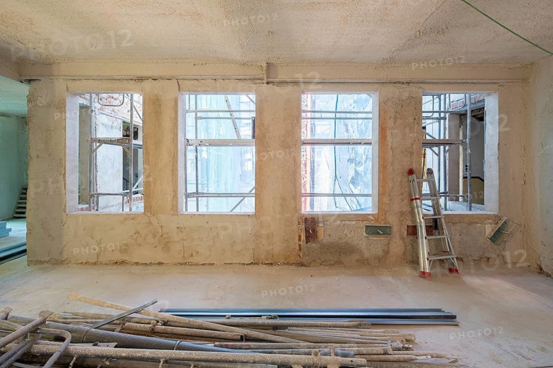 The interior of a building under construction, with large windows, scaffolding, and a metal staircase. The space is filled with building materials