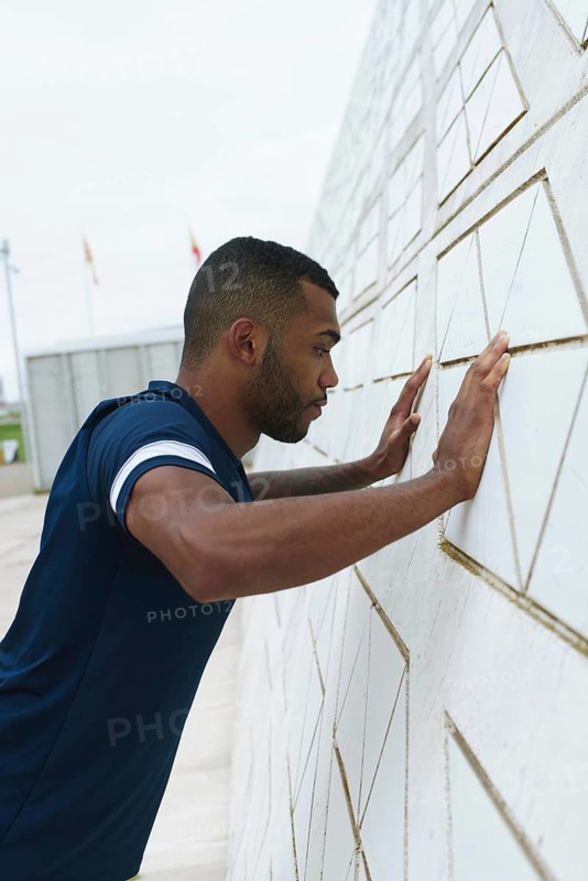 Black male athlete focuses intensely during an outdoor workout, pressing against a patterned wall. He embodies strength, determination, and perseverance in training