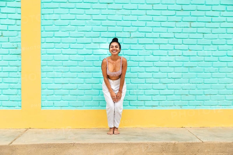A joyful young Latin woman poses against a colorful turquoise and yellow brick wall in the vibrant beach town of Mancora, Peru, symbolizing youthful energy and spirit