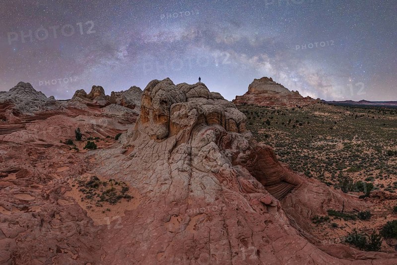 A distant, unrecognizable hiker stands atop a rugged rock formation under a star-filled sky in Utah, USA, highlighting the vast and wild beauty of the region