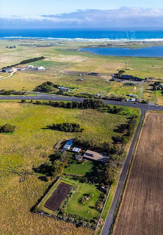 Aerial image showcases vast coastal farmland with a winding road, distant ocean, and cloudy sky in Victoria, Australia. Lush greenery, bright sky, and serene seascape create a tranquil scene
