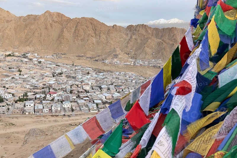 A stunning panorama of Leh, Ladakh, India, features vibrant Tibetan prayer flags fluttering in the foreground. Rugged mountains rise behind a bustling town below