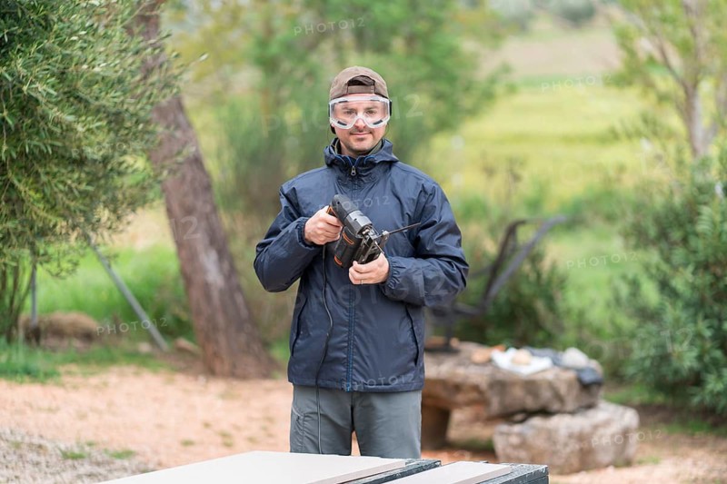 An happy man stands outdoors near a wooded area, holding a jigsaw for a carpentry project. He sports safety goggles, a cap, and a weatherproof jacket, focusing intently on his task