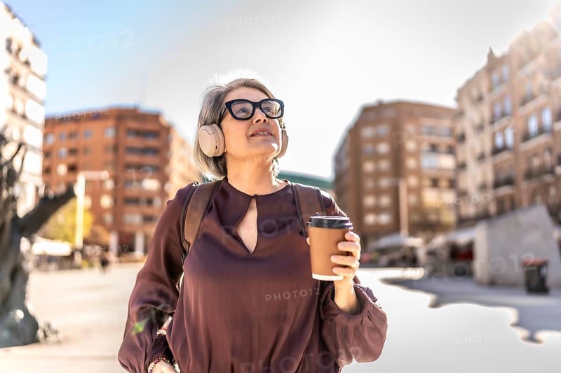 Mature woman with gray hair wearing headphones and elegant eyeglasses, holding a coffee cup, looking up enjoying the bright sun while walking outdoors through an urban city in daytime