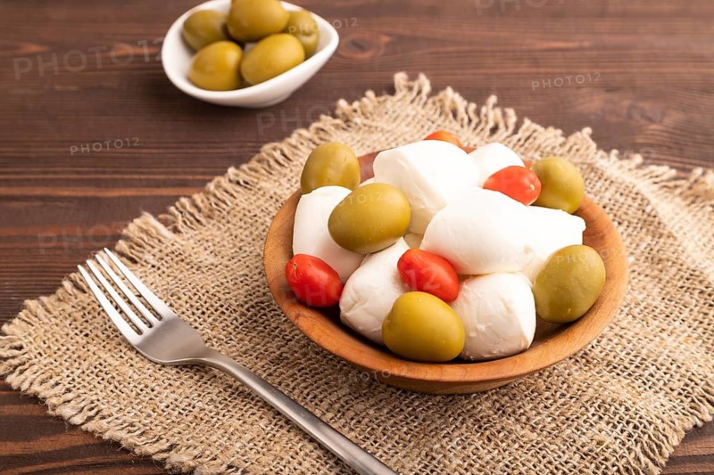 White Mozzarella cheese, with tomatoes and olives in wooden bowl on brown wooden background and linen textile, side view, close up
