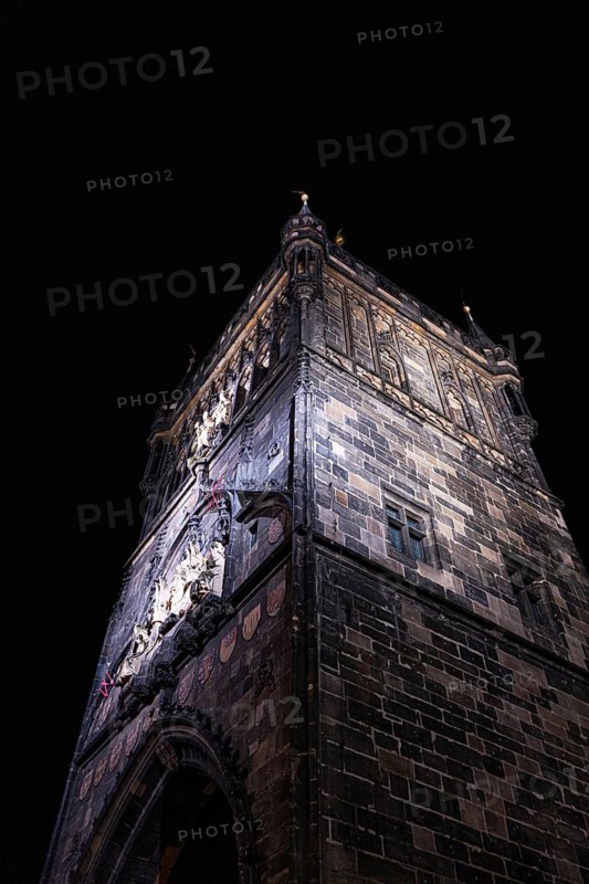A dramatic view of a Gothic tower in Prague, captured at night. The architectural details are highlighted by illumination against the dark sky, showcasing historical elegance