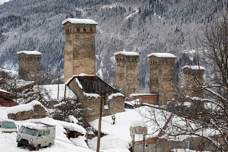 Stone watchtowers stand tall in a snow-covered village, nestled amidst mountainous terrain in Georgia Bare trees and rustic roofs complement the serene winter landscape
