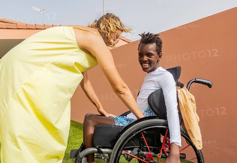 A person helps a boy in a wheelchair with cerebral palsy in pool. The scene depicts camaraderie and support under a clear sky, fostering a sense of inclusion and friendship
