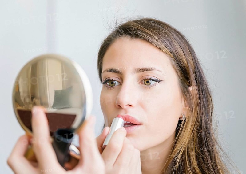 A woman carefully applies lipstick, focusing on her reflection in a handheld mirror. Her expression is intent, highlighting her natural beauty and makeup routine